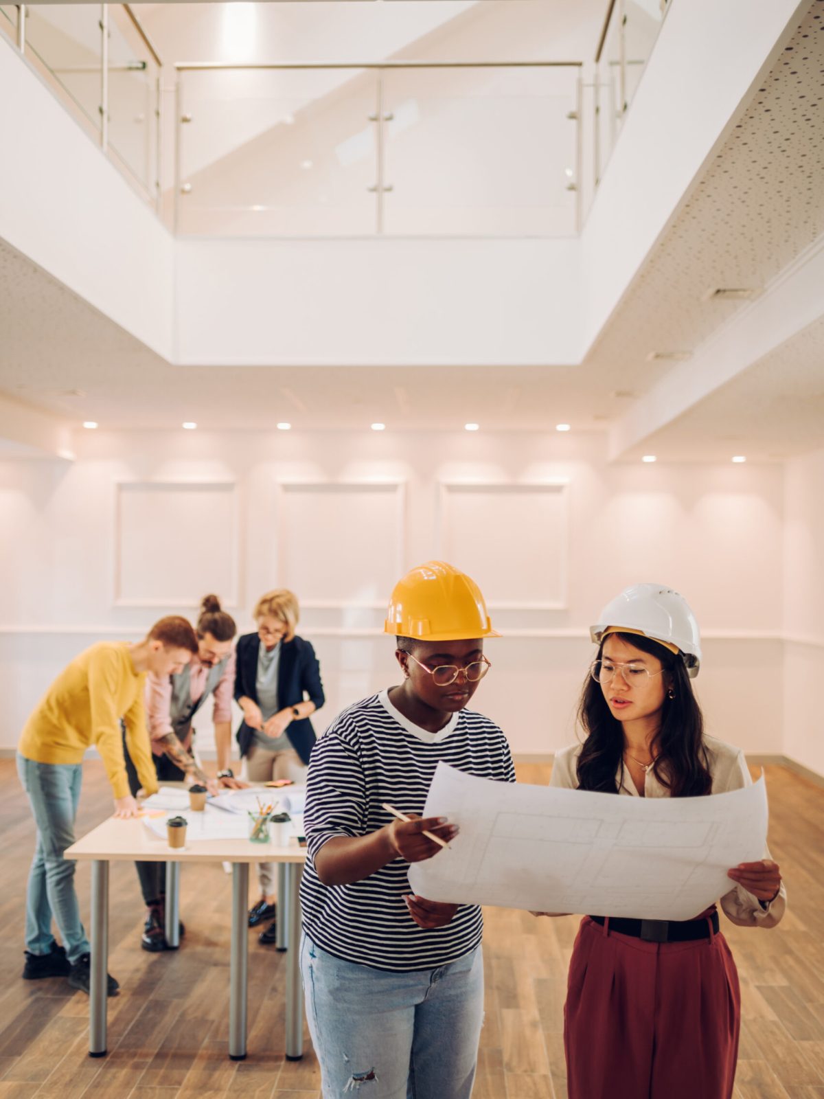 Two female architect looking at blueprint at architecture studio. Portrait of african american and vietnamese woman engineer in office with their colleagues having a meeting in the background.