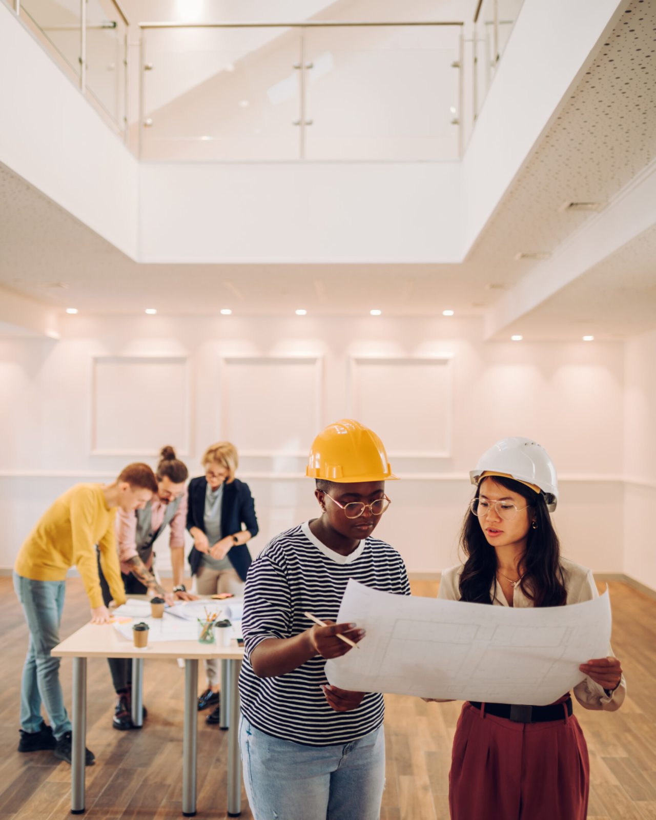 Two female architect looking at blueprint at architecture studio. Portrait of african american and vietnamese woman engineer in office with their colleagues having a meeting in the background.
